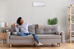 Woman relaxing on a couch under a wall-mounted AC unit, enjoying cool air and saving money.