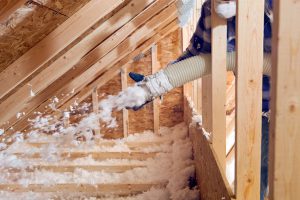 Worker blowing loose-fill insulation into attic space between wooden beams for energy efficiency.